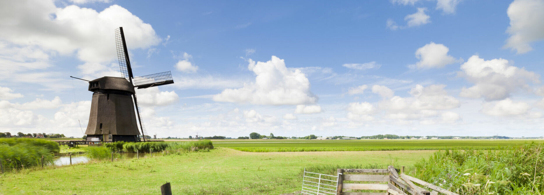 Een wei met een hek, een molen en een blauwe lucht met wolkjes.